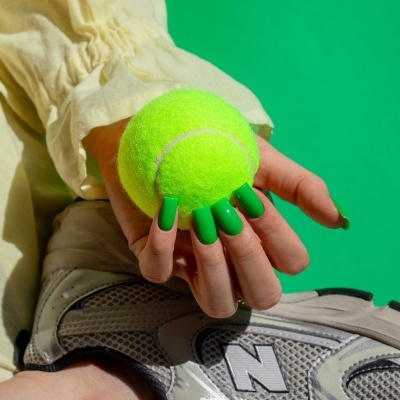 Mão segurando bola de ténis amarela com unhas verdes e tênis cinzento