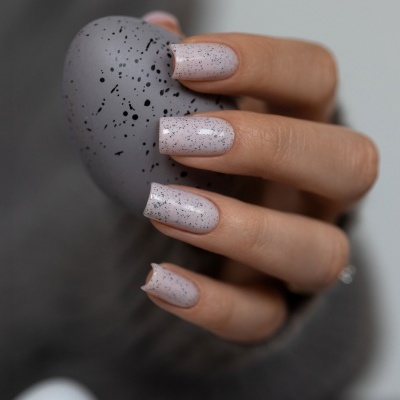 Mão com unhas pintadas de nude com manchas pretas, segurando objeto cinzento manchado.