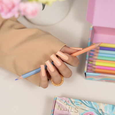 Mão com unhas postiças segurando lápis de cor azul claro junto a caixa de lápis coloridos pastel