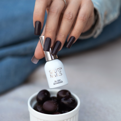 Frasco branco de verniz de unhas SAUTE NAILS em mão com unhas pintadas castanho escuro sobre fundo desfocado e taça branca com frutos escuros
