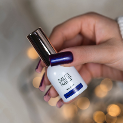 Frasco branco de verniz para unhas com tampa prateada segurado por mão com unhas roxas brilhantes