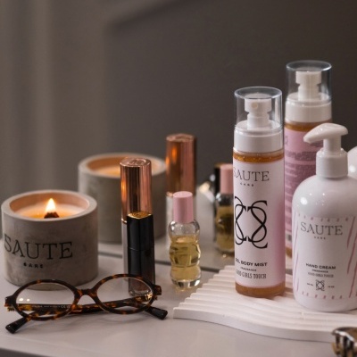 Produtos SAUTE CARE incluindo vela, gel body mist e creme mãos, com óculos castanhos sobre superfície branca junto a espelho.