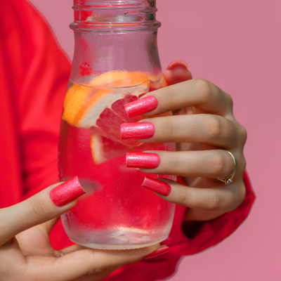 Mãos femininas com unhas longas cor de rosa metálico segurando frasco de vidro com bebida e frutas contra fundo rosa