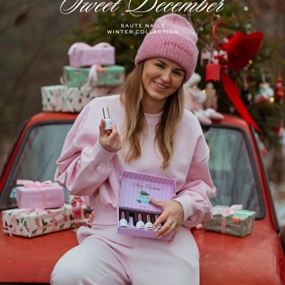 Mulher jovem com roupa rosa segurando caixa de esmaltes e esmalte, sentada em carro vermelho com presentes e árvore de Natal ao fundo.