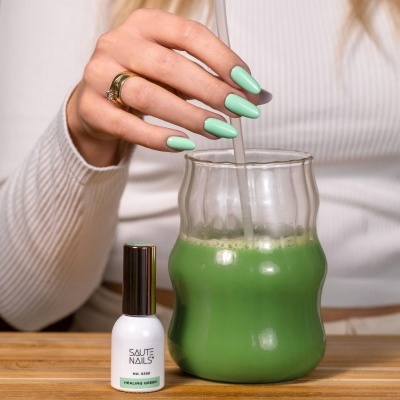 Frasco de verniz de unhas branco com tampa preta e unhas pintadas de verde claro junto a um copo com bebida verde.
