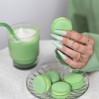 Mão com unhas verdes a segurar macaron verde ao lado de prato com mais macarons e bebida verde.