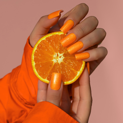 Mãos com unhas laranja cintilante segurando uma fatia de laranja com fundo rosa