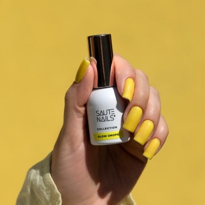 Frasco de verniz branco com tampa prateada em mão com unhas amarelas sobre fundo amarelo