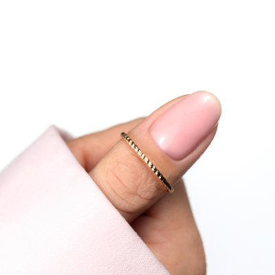 Anel dourado fino num dedo com unhas pintadas de rosa claro sobre fundo branco