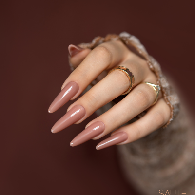 Mãos com unhas rosa nude e anéis dourados em fundo castanho