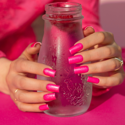 Mãos com unhas rosa brilhante segurando jarra de vidro com condensação sobre superfície rosa