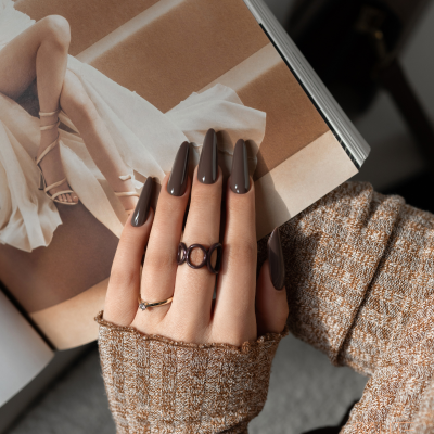 Mão feminina com unhas castanho escuro e anéis segurando revista aberta.