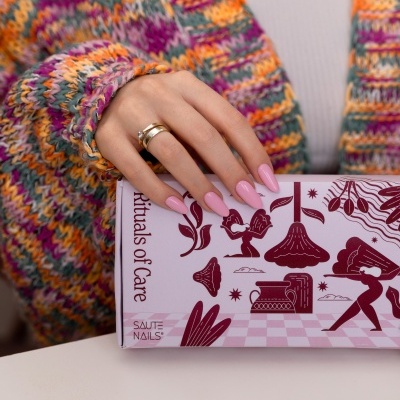 Mão com unhas rosa e anéis segurando caixa rosa com texto Rituals of Care