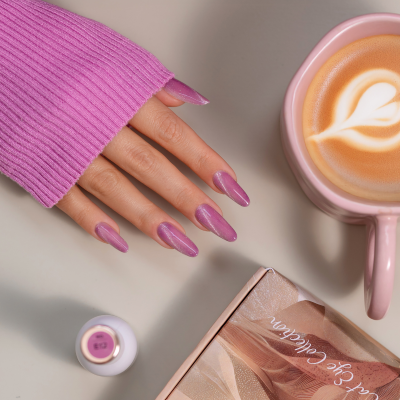 Mão com unhas pintadas de rosa metálico ao lado de frasco de verniz, caixa rotulada e chávena rosa com bebida decorada.