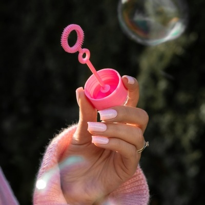 Mão com unhas brancas a segurar vareta cor-de-rosa para bolhas de sabão com uma bolha no ar e fundo desfocado.