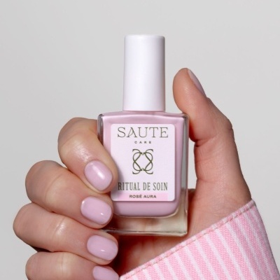 Frasco de verniz de unhas rosa claro com rótulo Saute Care Ritual de Soin Rosé Aura segurado por mão com unhas pintadas da mesma cor