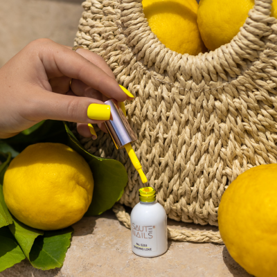 Mão a segurar pincel de verniz amarelo SAUTE NAILS junto a limões e mala de ráfia trançada.