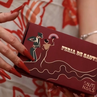 Caixa vinho com ilustração casal flamenco e texto FERIA DE SANTOS, segurada por mão com unhas vermelhas