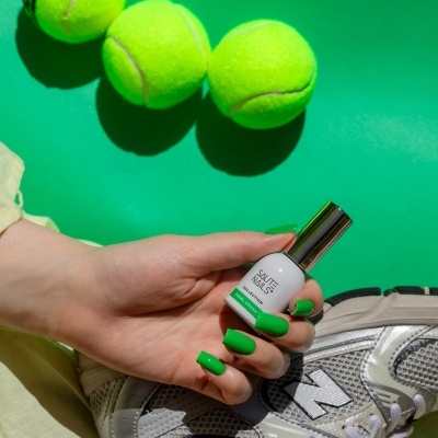 Mão com unhas verdes segurando verniz branco SAUTE NAILS perto de bolas de ténis e ténis cinza