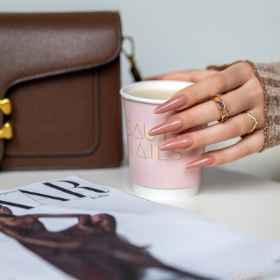 Mão com unhas castanho nude segurando copo rosa com letras douradas, mala castanha de couro e revista de moda em fundo branco.