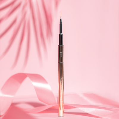 Pincel de unhas metalizado dourado e preto com letras SAUTE NAILS sobre fundo rosa e fita de cetim.