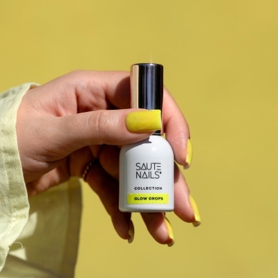 Frasco branco de verniz SAUTE NAILS COLLECTION GLOW DROPS na mão com unhas amarelas