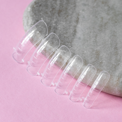 Unhas postiças transparentes em semi-círculo sobre base cinzenta e fundo rosa