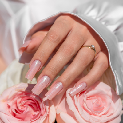 Mão feminina com unhas rosa cintilantes segurando rosas cor-de-rosa e anel dourado.