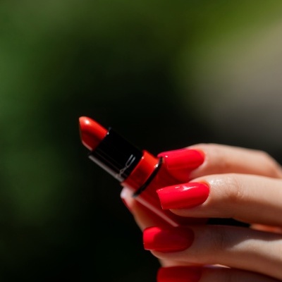 Batom vermelho vivo MAC numa mão com unhas vermelhas, fundo verde desfocado