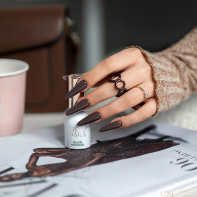 Mão com unhas castanho escuro segurando frasco de verniz de unhas branco SAUTE NAILS sobre revista