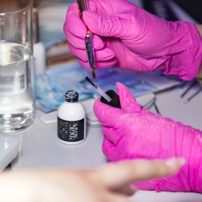 Mãos com luvas cor-de-rosa segurando pincel e frasco de produto para unhas em mesa