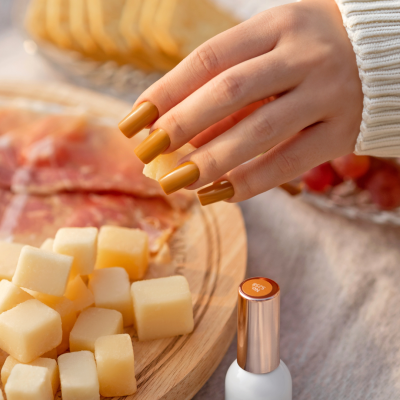 Mão com unhas pintadas de castanho mostarda junto a tábua de queijo, presunto e verniz SAUTÉ NAILS