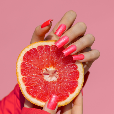 Mãos com unhas cor-de-rosa metálico segurando meio toranja vermelha em fundo rosa