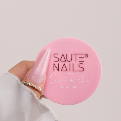 Frasco rosa de cosmético para unhas SAUTE NAILS com texto One Touch Glitter Pink, mão com unhas rosa gliter segurando.
