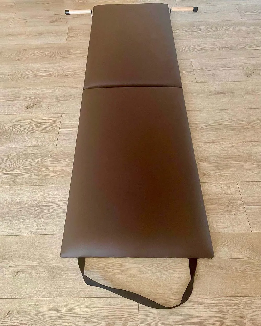 Colchonete de exercício dobrável castanho com alça preta num chão de madeira clara.