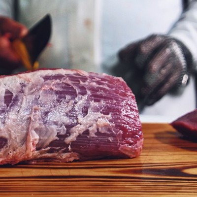 Pedaços de carne crua vermelha em tábua de madeira com pessoa ao fundo vestindo luva metálica e camisa branca