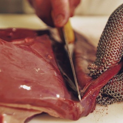 Pessoa a cortar carne crua com faca e luva de malha metálica.
