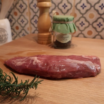 Pedaço de carne crua numa bancada de madeira com alecrim, frasco com tampa verde, saleiro e azulejos coloridos ao fundo