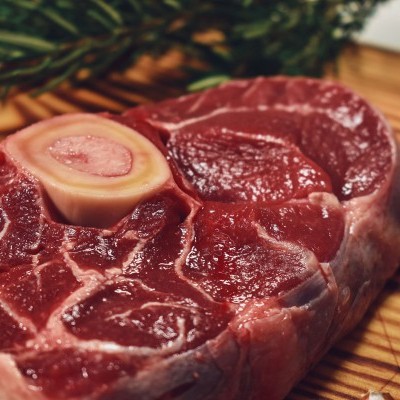 Pedaço cru de carne vermelha com osso no centro sobre tábua de madeira com ervas ao fundo