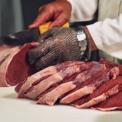 Pessoa corta peça grande de carne vermelha em fatias com cutelo, usando luva metálica na mão.