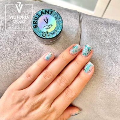 Mão com unhas pintadas com gel brilhoso verde e azul ao lado de frasco Victoria Vynn em fundo cinzento