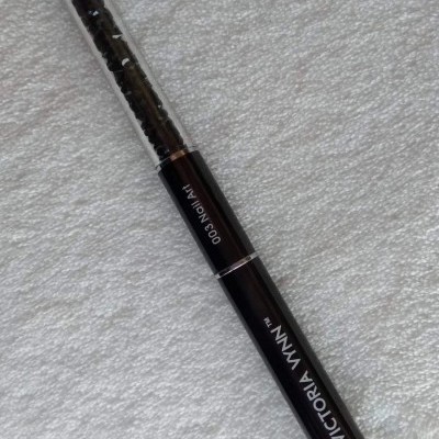 Lápis para olhos preto com tampa transparente e texto visível, sobre tecido branco.