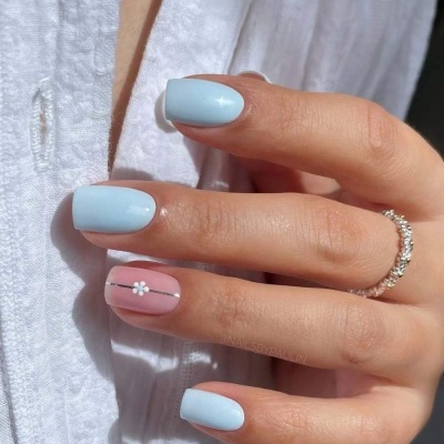 Unhas pintadas em azul claro e rosa com flor branca e riscas pretas em mão feminina com anel sobre tecido branco.