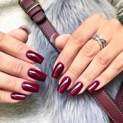 Unhas longas vermelhas escuras brilhantes com anéis em prata sobre tecido cinza e bolsa vinho