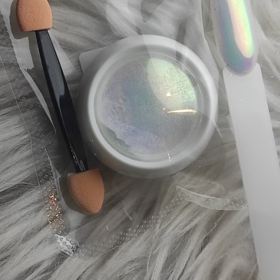 Pequena embalagem branca com pó cintilante iridescente e aplicador de esponja dupla preta sobre tecido peludo cinzento