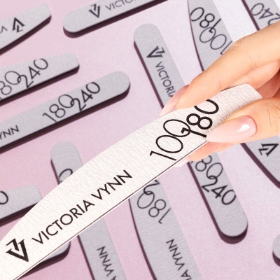 Lixas para unhas cinza com texto preto VICTORIA VYNN e números de granulação, sobre fundo rosa, com mão a segurar uma delas.