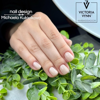 Mão com unhas nude brilhantes sobre folhas verdes artificiais, com texto nail design e logotipo Victoria Vynn