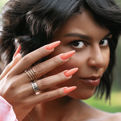 Mulher com unhas laranja longas e anéis prateados, fundo verde ao ar livre