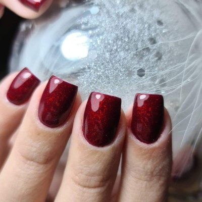 Unhas vermelhas brilhantes com gel segurando uma esfera prateada decorativa com penas brancas.