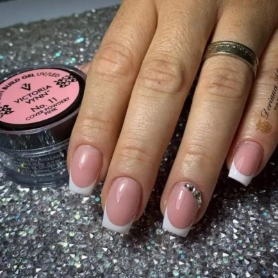 Unhas postiças estilo francesa em mãos femininas com decoração e frasco de gel Victoria ao lado.